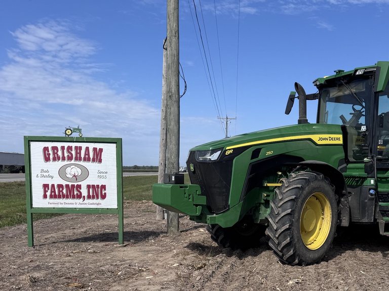 The Grisham Farms, Inc. sign beside a green tractor on bare farm ground under a blue sky.