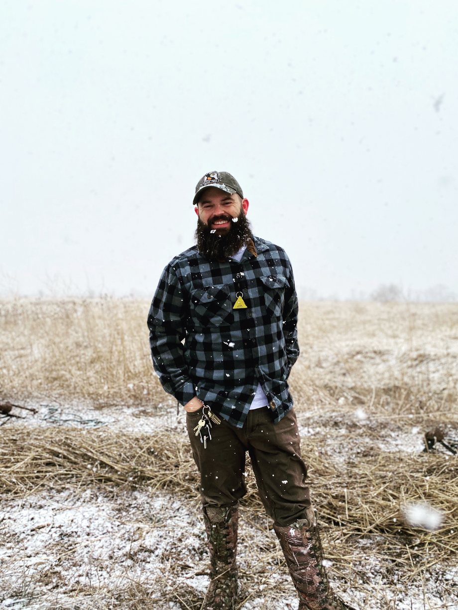 Blake Grisham smiles in falling snow in a grassy field, in a plaid flannel shirt and a cap.