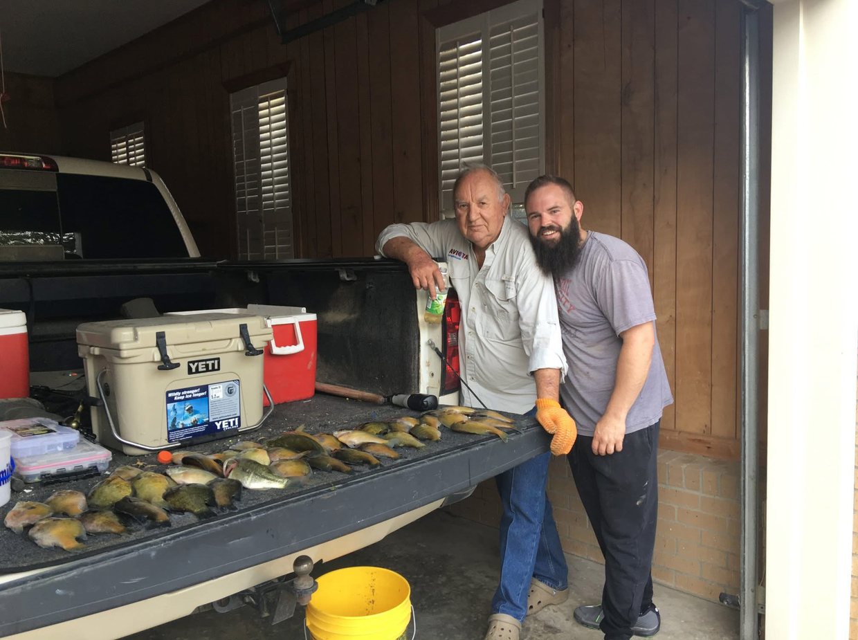 Daddy Bob and Blake stand at the open tailgate of a pickup in a garage, rows of sunfish and one bass laid across the tailgate, coolers beside them and a yellow bucket on the floor below.