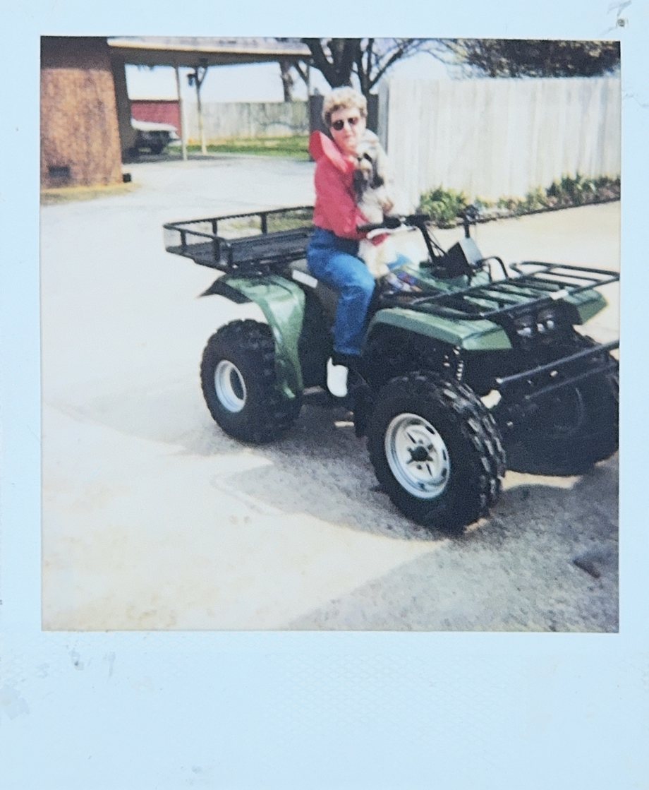 An instant photograph: a woman with short curly hair, sunglasses, a red jacket, and jeans sits on a green four-wheeler with black cargo racks, holding a small long-haired dog against her chest. The four-wheeler is parked on a concrete drive, with a brick wall, a carport, and a white wooden fence behind it.